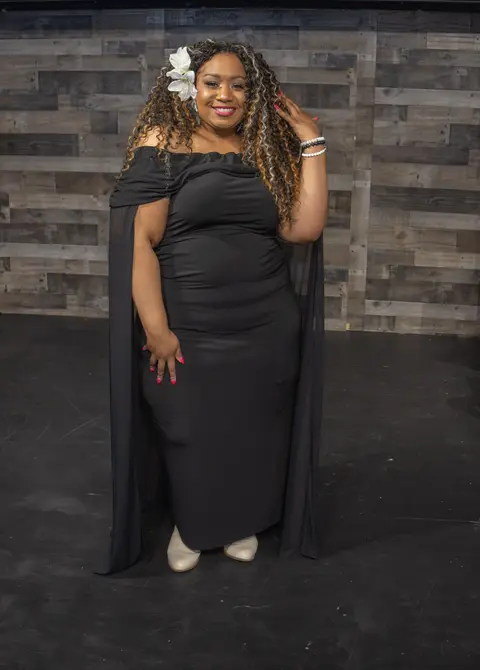 Andrea “Vocab” Sanderson full-length in a black caped gown with a white flower in her hair, against a wood-panel wall