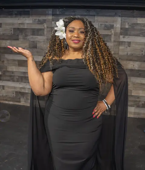 Andrea “Vocab” Sanderson smiling in a black caped gown, one hand raised, white flower in her hair